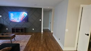 A modern living room with a flat-screen TV mounted on a textured wall, wooden flooring, and a door leading to another room. - 10 mins from Mercedes Benz stadium! (Atlanta)