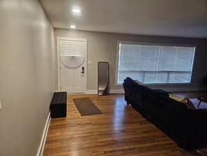 Um quarto com piso de madeira, um sofá preto, um banco, uma porta com um detalhe de leque e uma grande janela com persianas.