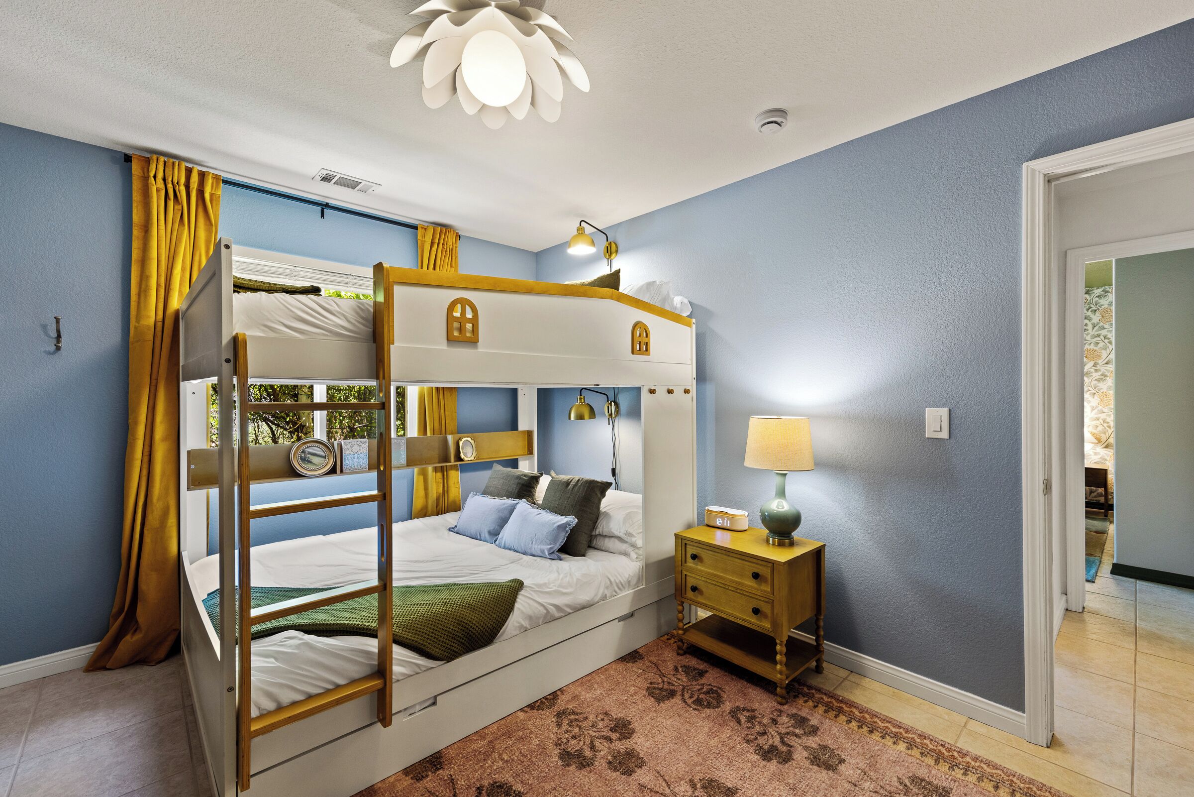 A bunk bed room with a window, a bedside table with a lamp, and a door to another room.