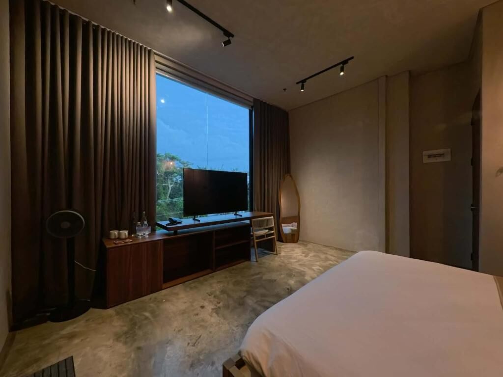Une chambre d’hôtel moderne avec un grand lit, un téléviseur posé sur un meuble en bois, un ventilateur et une vue sur l’extérieur.