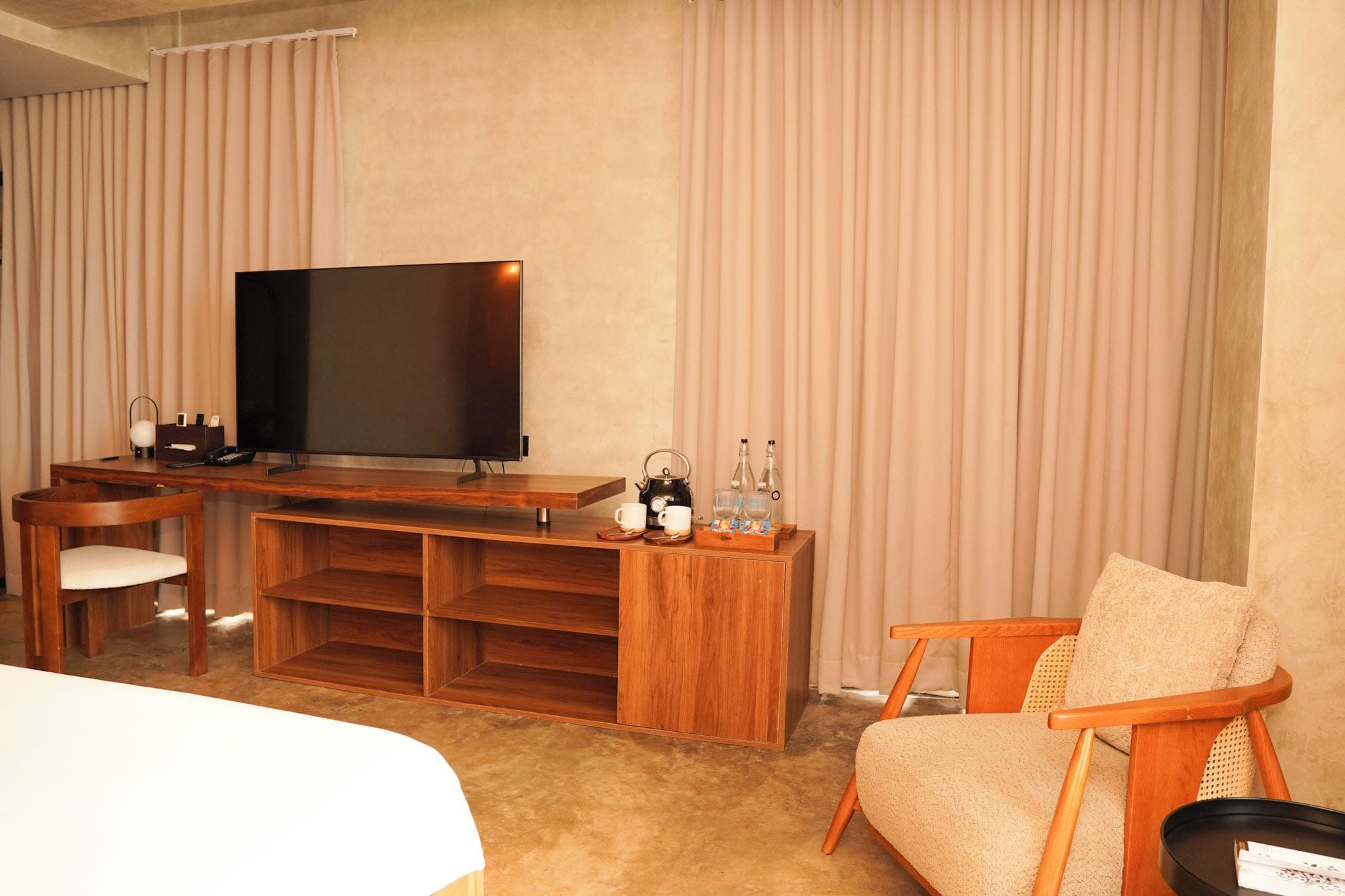 Quarto de hotel com televisão de tela plana, mobiliário de madeira, sofá e escrivaninha com chaleira e xícaras.