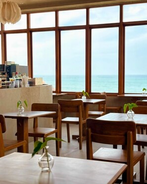 A restaurant with wooden tables and chairs, a bar area, and large windows offering a view of the sea. - The Salt Boutique Hotel by Wyns (San Fernando)