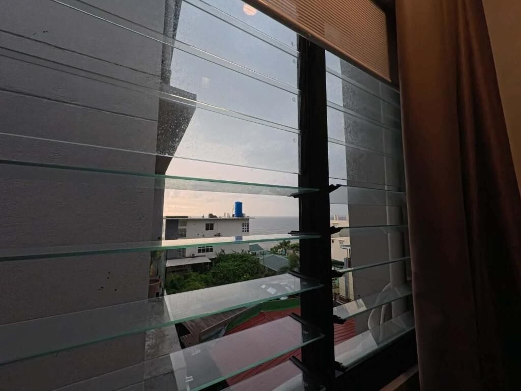 A view from a window with horizontal blinds, a curtain partially visible, and a glimpse of buildings and greenery outside.