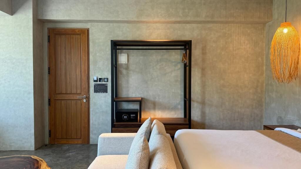 A hotel room with a wooden door, a bed with white bedding, a small bedside table, a wall-mounted heater, and a hanging light fixture.