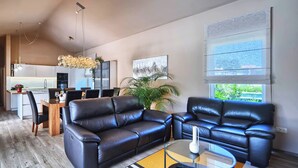 Ein modernes Wohnzimmer mit Ledersofas, einem Glastisch und einem Essbereich, im Hintergrund ist eine Küche zu sehen.