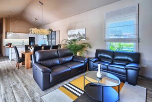 Ein modernes Wohnzimmer mit Ledersofas, einem Glastisch und einem Essbereich, im Hintergrund ist eine Küche zu sehen.
