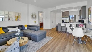 Uma sala de estar moderna com um sofá em formato de L, uma mesa de centro e uma área de jantar com cadeiras brancas.