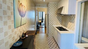 A narrow hallway leading to a kitchen with a gas stove, a dining area with a table and chairs, and a wall with a floral painting.