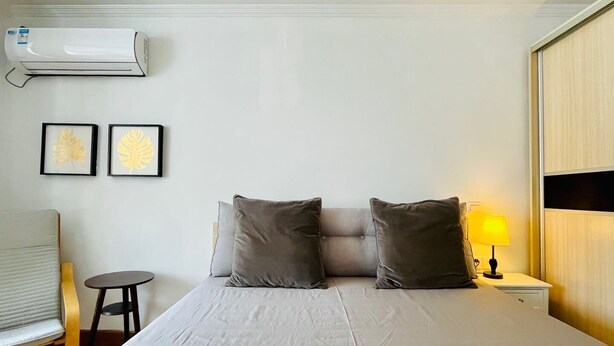 A bedroom with a bed, two pillows, a nightstand with a lamp, a chair, and an air conditioning unit on the wall.