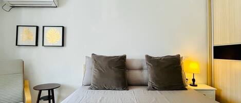 A bedroom with a bed, two pillows, a nightstand with a lamp, a chair, and an air conditioning unit on the wall.