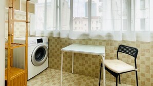 A laundry room with a washing machine, a table, a chair, and a wooden shelf unit.