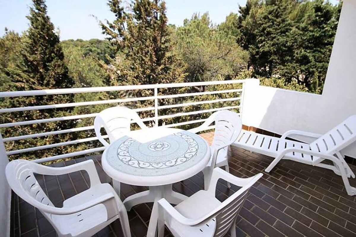 Ein Balkon mit weißen Plastikstühlen und einem Tisch, umgeben von einem weißen Geländer, mit Blick auf einen Garten mit Bäumen.