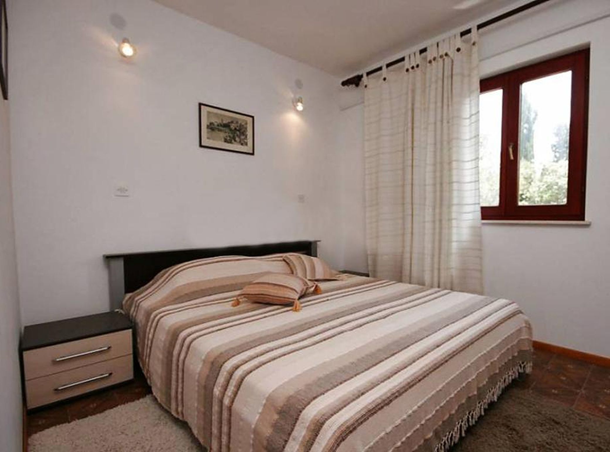 Une chambre à coucher comprenant un lit, une table de chevet, une fenêtre avec des rideaux et un luminaire fixé au mur.