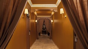 A corridor with a marble floor, flanked by curtains and wall sconces, leading to a room with a bed and a desk.