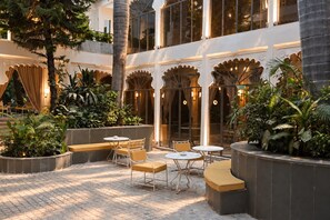 An outdoor seating area with tables and benches, surrounded by lush greenery and architectural details. - Mewar Manor by My Downtown Hotels - Jhadol Haveli (Udaipur)