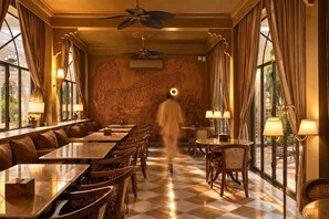 A restaurant with a checkered floor, long tables, and chairs, with a large stone wall and windows with curtains. - Mewar Manor by My Downtown Hotels - Jhadol Haveli (Udaipur)