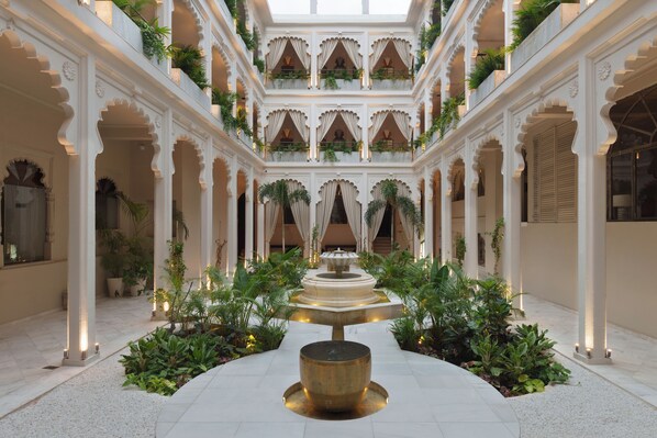 A spacious atrium with a central fountain, surrounded by lush greenery and white architectural details. - Mewar Manor by My Downtown Hotels - Jhadol Haveli (Udaipur)