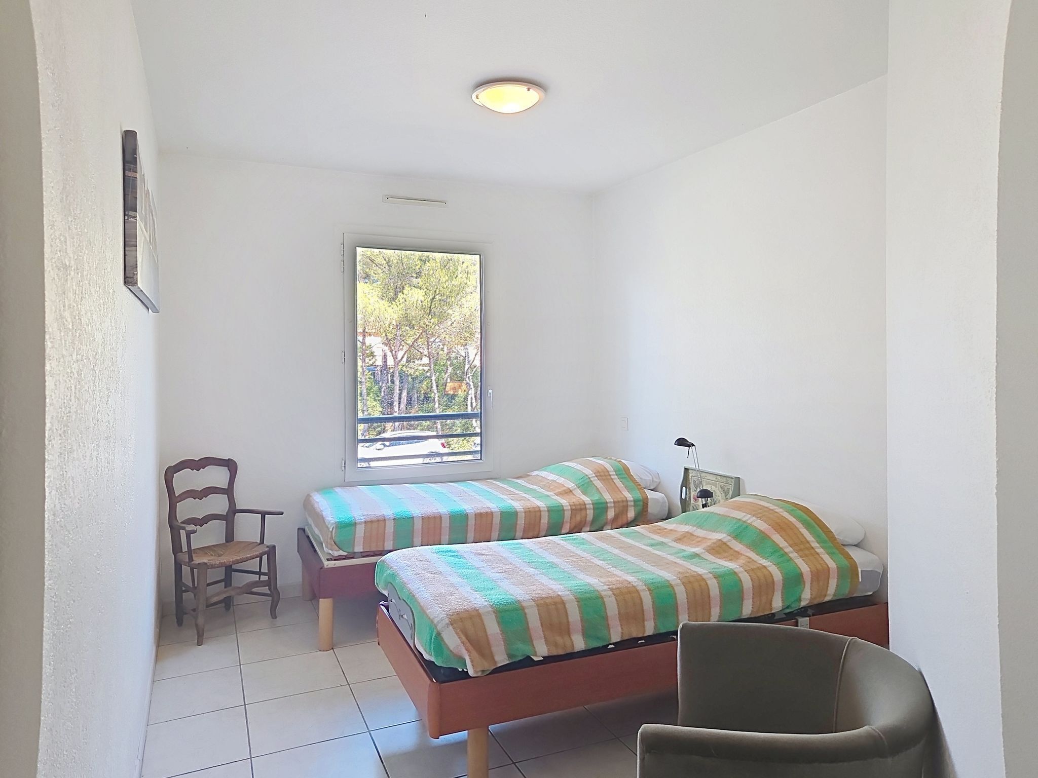 Una habitación pequeña y limpia con dos camas individuales, una silla, un ventanal con vistas a árboles y una lámpara en el techo.