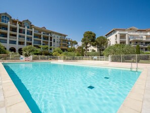 A swimming pool with clear blue water, surrounded by a tiled deck. In the background, there are multi-story buildings with balconies and a fenced garden area. - Le Grand Parc by Interhome (Saint Aygulf)