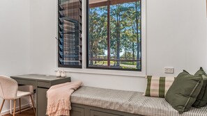 A room with a desk, a bench with cushions, a large window with a view of trees and a fence, and a power outlet on the wall.