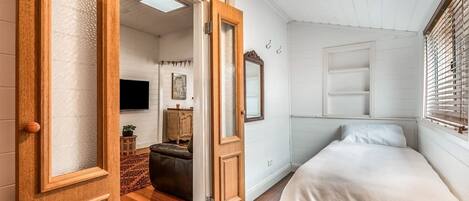 Um quarto com uma cama, cômoda, televisão e porta que dá para outro espaço.
