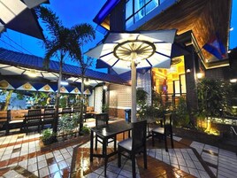 Un patio al aire libre con mesas y sillas, un paraguas grande y un edificio con techo de tejas.