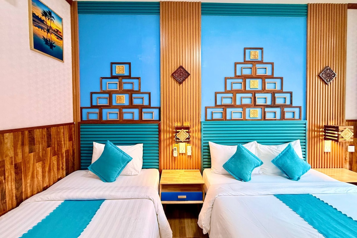 Habitación de hotel con dos camas, cabecero de madera, pared azul con patrones geométricos y una pintura de un atardecer.