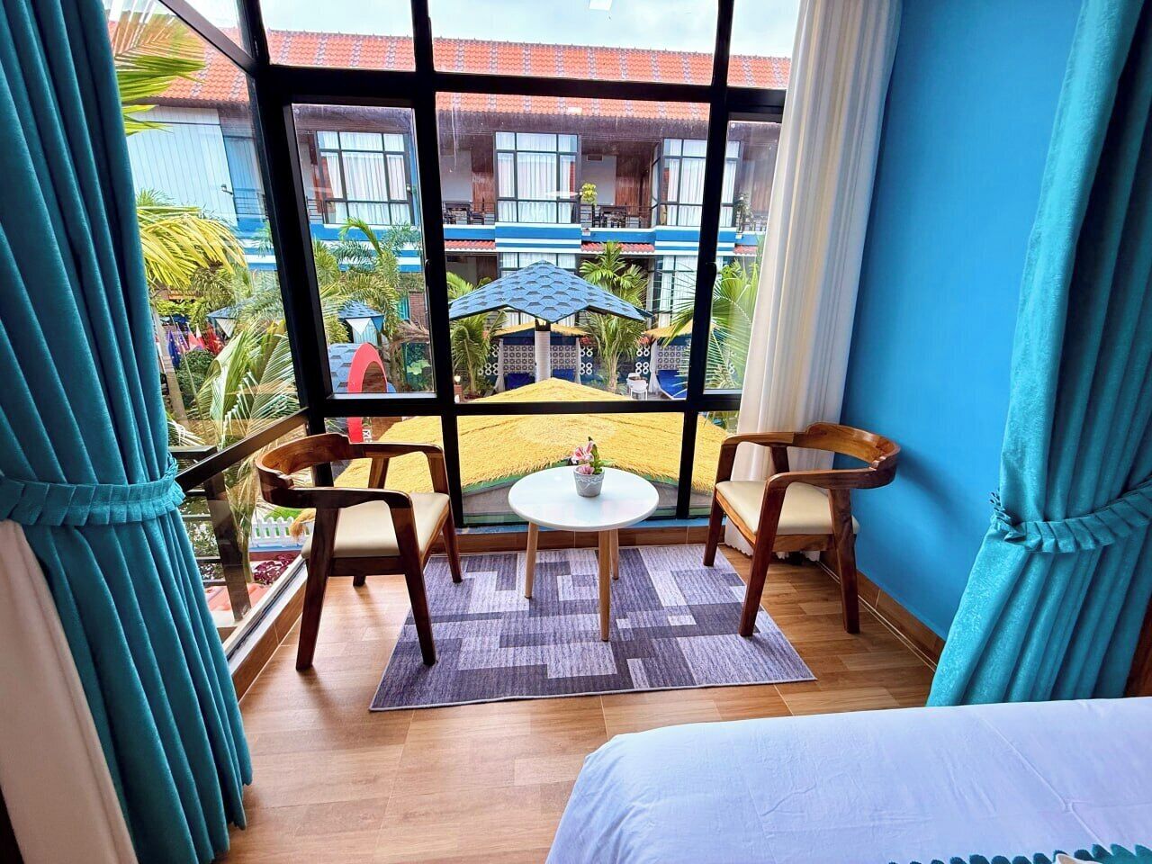 Habitación con cama, una mesita, dos sillas y vista a un edificio con techo azul y palmeras.