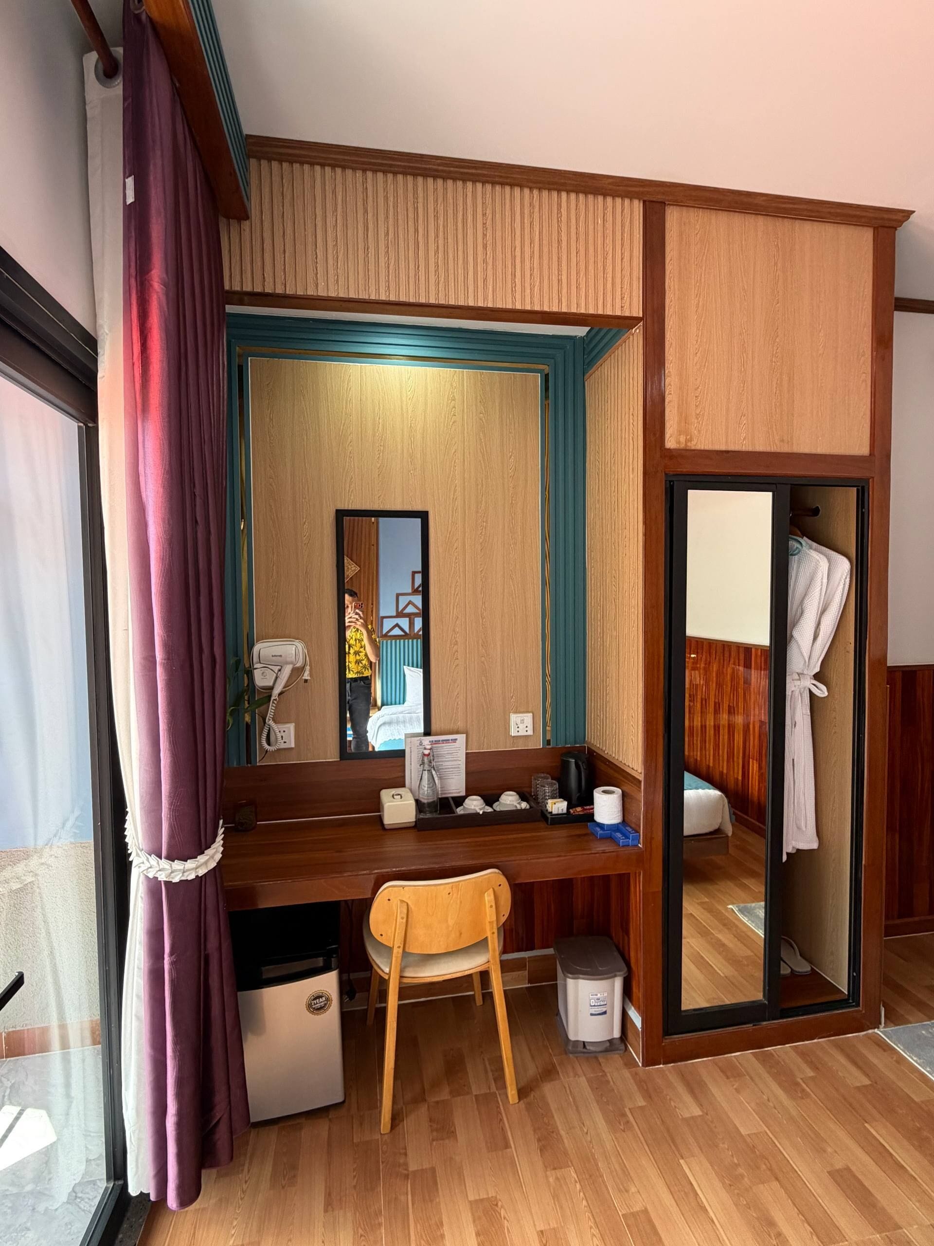 A compact hotel room with a wooden desk, a chair, a mirror, a small trash bin, a robe hanging, and a wooden wardrobe.