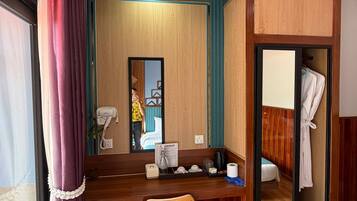 A compact hotel room with a wooden desk, a chair, a mirror, a small trash bin, a robe hanging, and a wooden wardrobe.