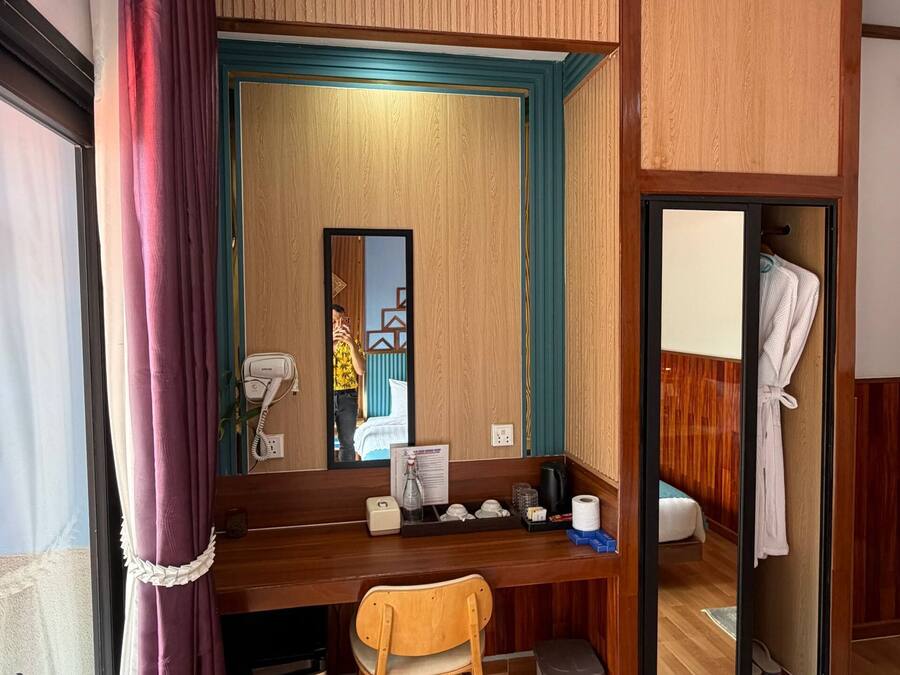 A compact hotel room with a wooden desk, a chair, a mirror, a small trash bin, a robe hanging, and a wooden wardrobe.