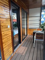 Exterior de madera con puerta corredera de vidrio, una mesita con una silla y una ventana con persianas.