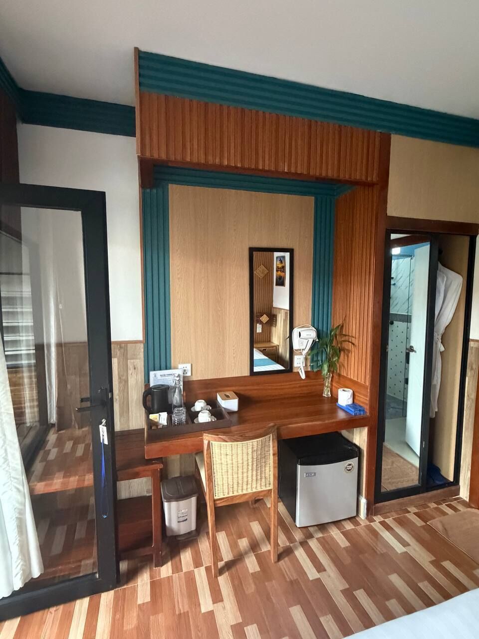 A hotel room with a wooden desk, a chair, a small trash bin, a mirror, and a plant.