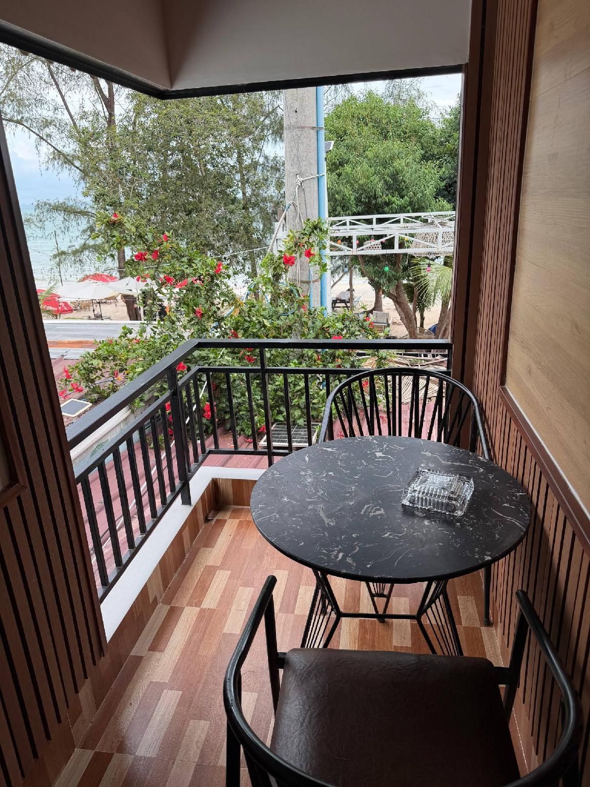 A balcony with a round table and chairs, a view of a garden with red flowers, and a wooden floor.
