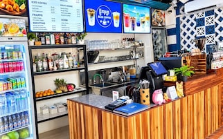 Interior de una cafetería con mostrador de madera, estanterías llenas de bebidas, una pizarra con el menú y un refrigerador con refrescos.