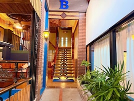 Entrada de un hotel con una escalera que lleva al interior, un letrero con la letra 'B' y una planta en maceta a la derecha.