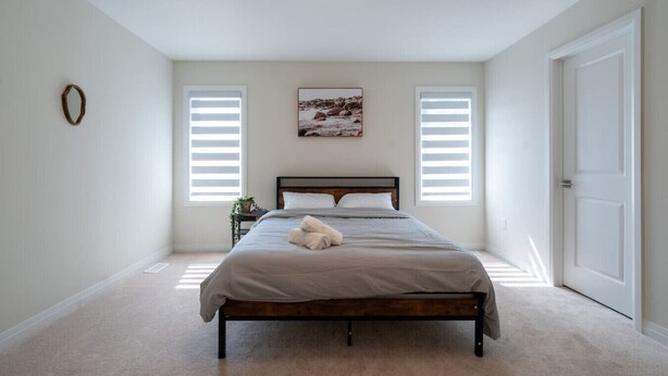 A bedroom with a bed, two windows with blinds, a door, and a ceiling light. - Spacious Modern Orillia Family Retreat- Hot Tub (Severn)