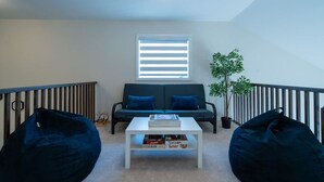 A loft-style room with a sofa, two bean bags, a coffee table with books, a potted plant, and a window with blinds. - Spacious Modern Orillia Family Retreat- Hot Tub (Severn)
