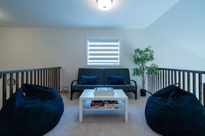 Ett rum i loftstil med en soffa, två sittpuffar, ett soffbord med böcker, en krukväxt och ett fönster med persienner.