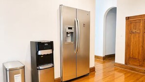 Uma geladeira de aço inoxidável com dispensador de água, uma lixeira e um armário de madeira em um quarto com piso de madeira.
