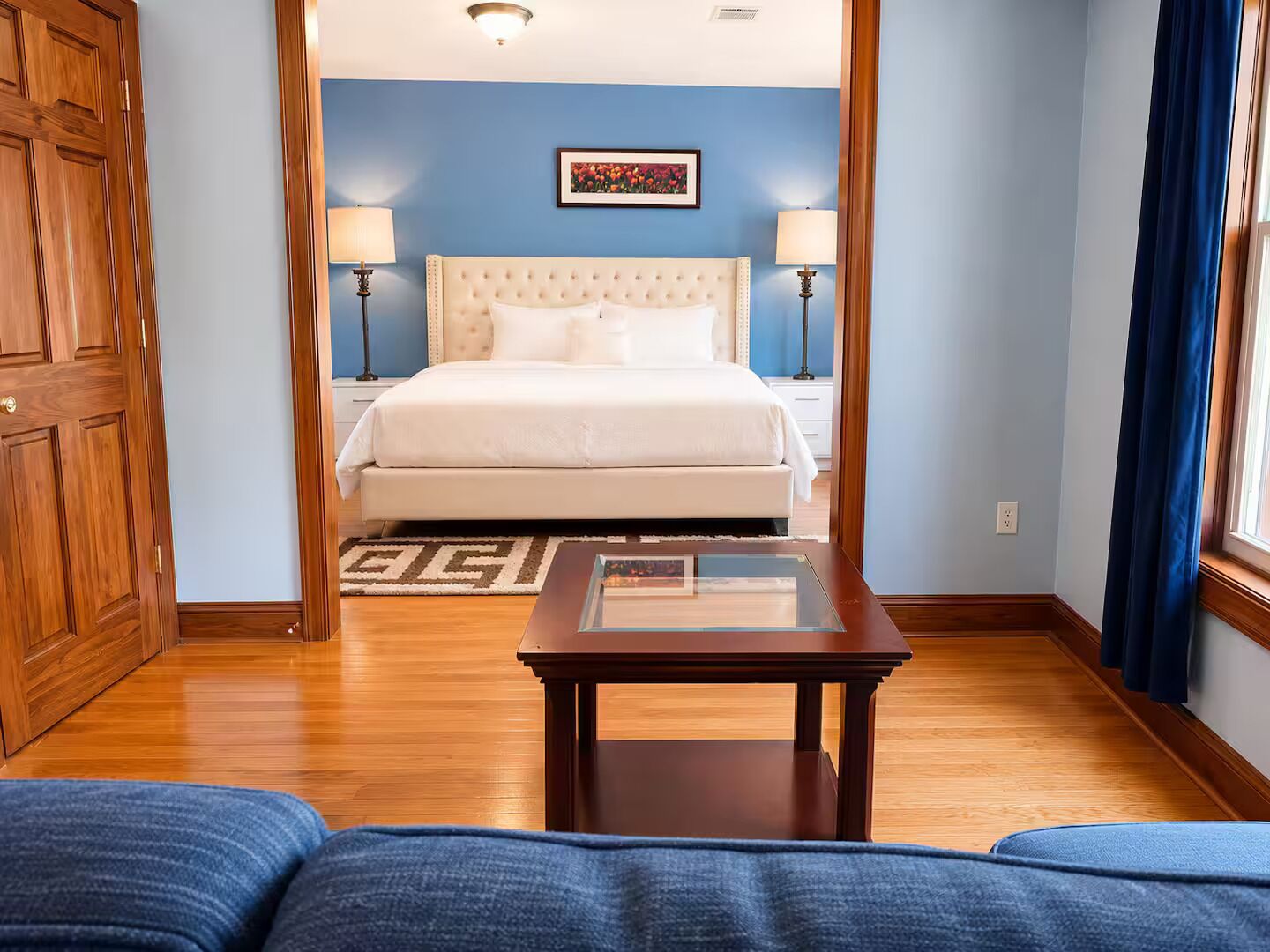 Una camera d'albergo con un letto, lampade sui comodini, un pavimento in legno e vista attraverso una porta aperta.