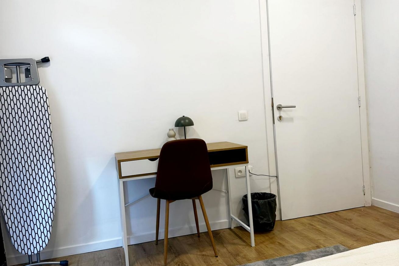 A room with a wooden desk, a red chair, a white door, and a patterned ironing board.