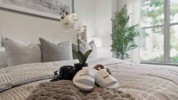 A bedroom with a bed, pillows, a plant, a pair of slippers, and a framed picture on the wall.