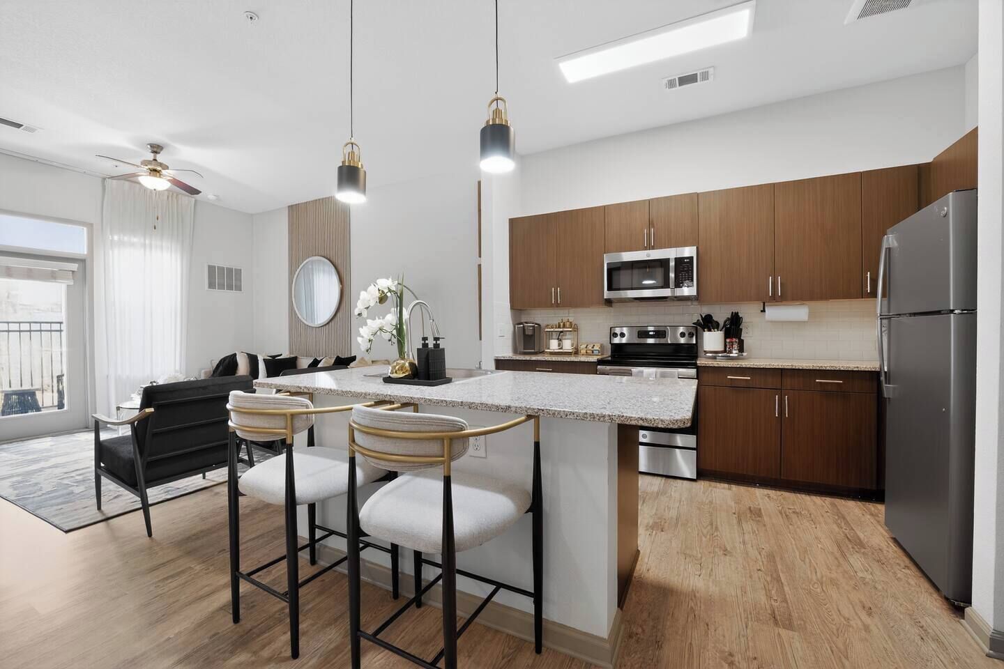 A modern kitchen with a central island, bar stools, wooden cabinets, stainless steel appliances, and a ceiling fan.