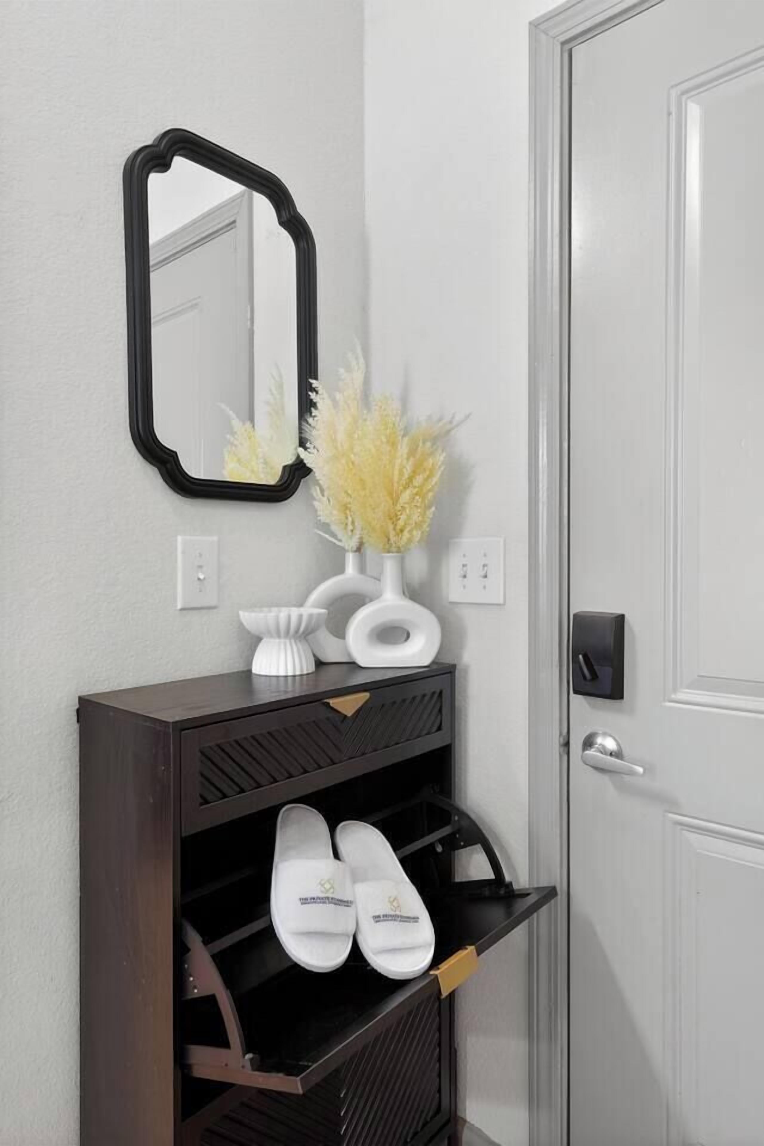 A hallway with a dark wooden console table, a white vase with yellow flowers, a black-framed mirror, and a white door.