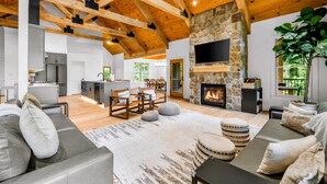 A spacious living area with a stone fireplace, wooden beams, and a modern kitchen in the background. - Lake Anna Lakefront | Beach, Game Room, Hot Tub (Mineral)