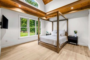 Ein Raum mit einem Himmelbett, großen Fenstern, Holzbalkendecke und einem kleinen Nachttisch mit Pflanze.