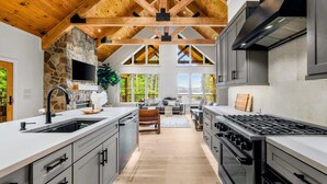 A modern kitchen with a stone fireplace, wooden ceiling beams, and a large island. - Lake Anna Lakefront | Beach, Game Room, Hot Tub (Mineral)