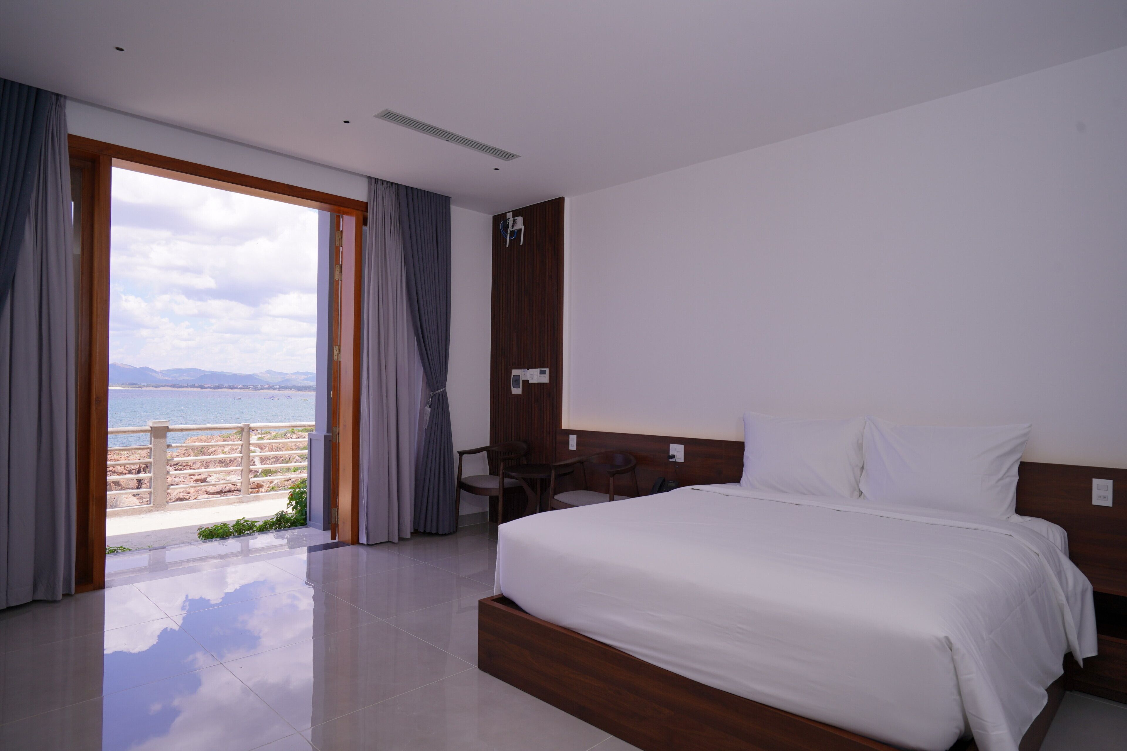 Habitación de hotel con una cama grande, cabecero de madera, un escritorio y vista al mar a través de un ventanal.