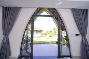 A room with a large glass door leading to an outdoor area with a thatched-roof structure and a lawn. - Blue Haven Resort Quy Nhon (Xuan Loc)